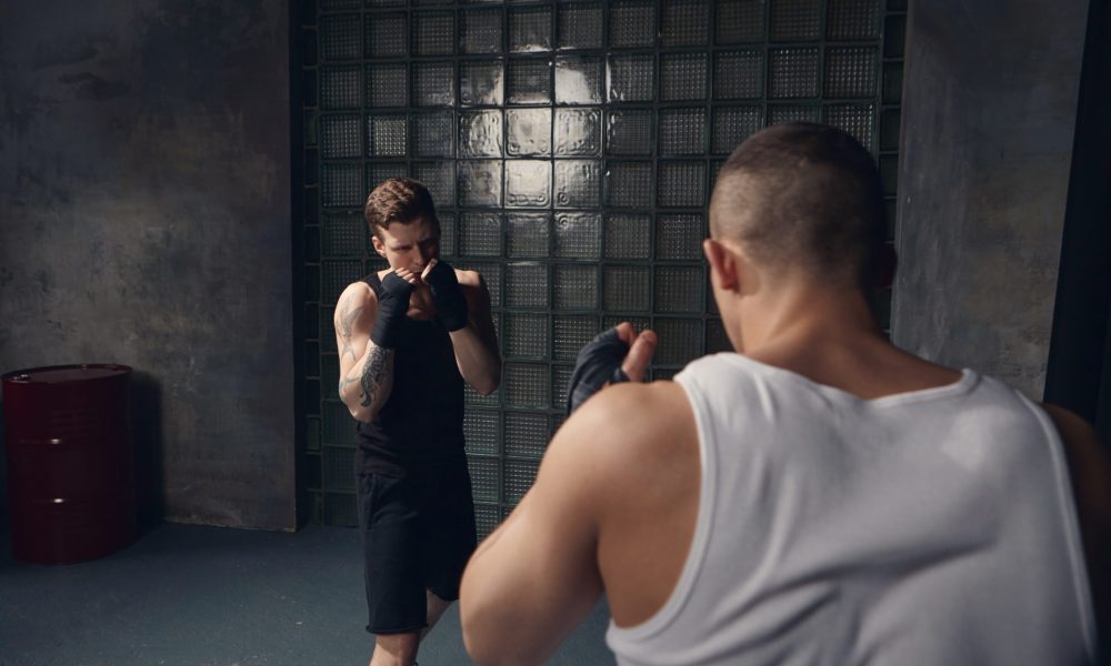 Back view of unrecognizable male fighter with broad muscular shoulders fighting against young Caucasian guy with tattoos wearing black outfit and bandages. Sports, martial arts and competition