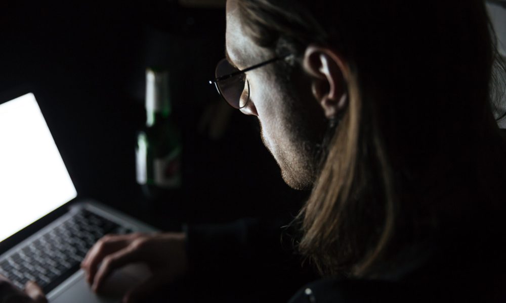 Photo of young concentrated man using laptop computer at home indoors at night. Looking at laptop display.